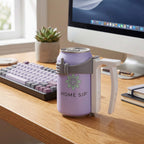 A lavender can-shaped insulated mug with a white handle and gray grip, shown on a wooden desk in front of a computer monitor. The mug has a green flower logo and the text 'HOME SIP'.