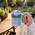 A hand holds a can-shaped tumbler with a mint green handle and matching grip in an outdoor backyard setting. The can has a white label with a green flower logo and the text 'HOME SIP'.