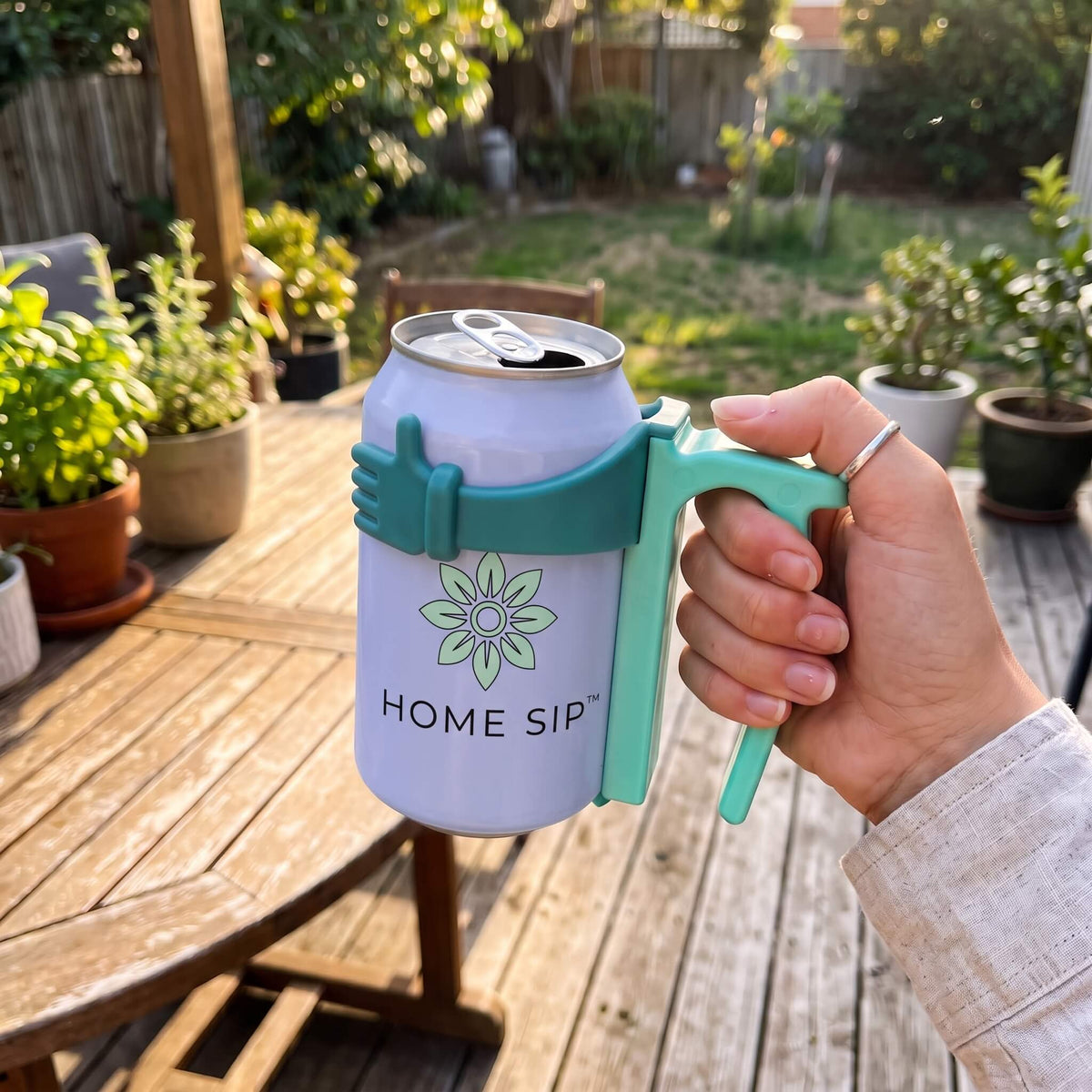 A hand holds a can-shaped tumbler with a mint green handle and matching grip in an outdoor backyard setting. The can has a white label with a green flower logo and the text 'HOME SIP'.